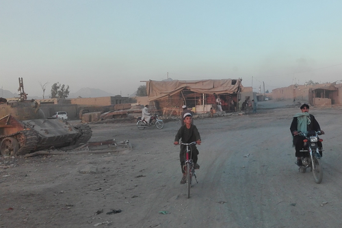 Ein Kind auf einem Fahrrad und ein Mann auf einem Moped in einem afghanischen Dorf bei Kandahar, links ist ein Panzer zu sehen. 