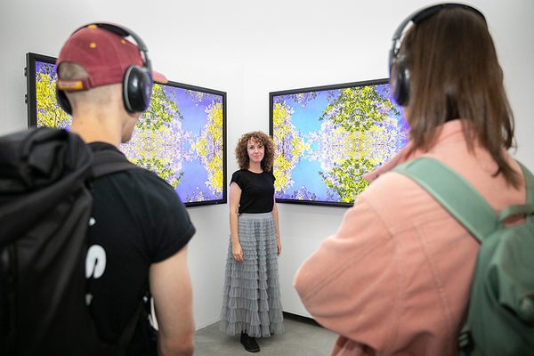 Zwei Besucherinnen tragen Kopfhörer und schauen sich auf zwei Bildschirmen eine Kunstinstallation an. Zwischen den Bildschirmen steht eine Frau. 