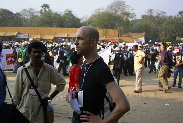 Jochen Steinhilber steht auf dem Gelände des Weltsozialforums 2004 in Mumbai. Weitere Besucher:innen gehen über das Gelände. Es sind Informationsstände von verschiedenen Organisationen aufgebaut. 