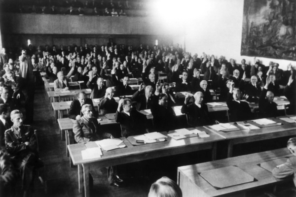 Schwarz-Weiß Fotografie von: Abstimmung über das Grundgesetz im Parlamentarischen Rat am 08.05.1949. Zu sehene sind viele sitzende Personen, die teilweise die Hand heben und alle nach links schauen.