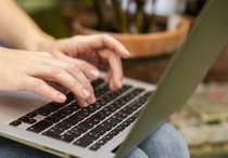 Eine Person schreibt etwas auf ihrem Laptop auf ihrem Schoß. Es ist nur der Laptop und ihre Hände zu sehen.