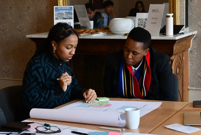 Zwei Teilnehmer:innen des GenSouth-Programms 2026 bei der Gruppenarbeit. Plakate liegen auf dem Tisch vor ihnen.