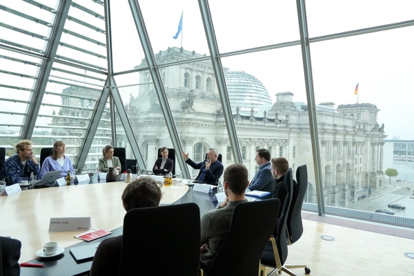 Die Teilnehmenden sitzen an einem Tisch mit Michael Roth, im Hintergrund das Bundestagsgebäude mit der Deutschlandflagge und der EU-Flagge.