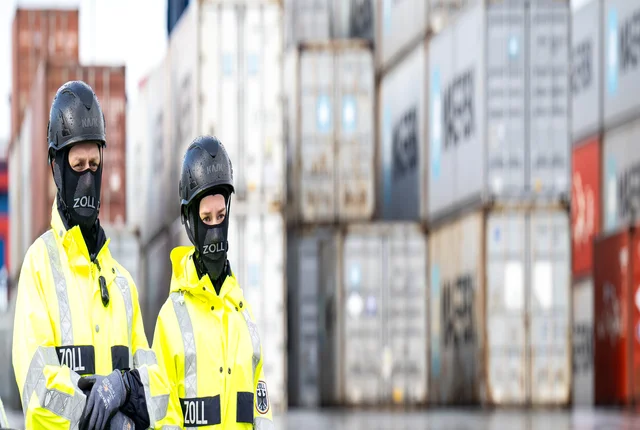Zwei Polizist_innen stehen in einem Hafen vor Containern.