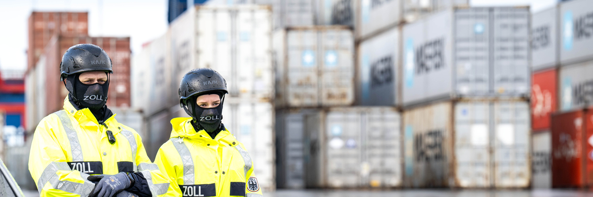 Zwei Polizist_innen stehen in einem Hafen vor Containern.