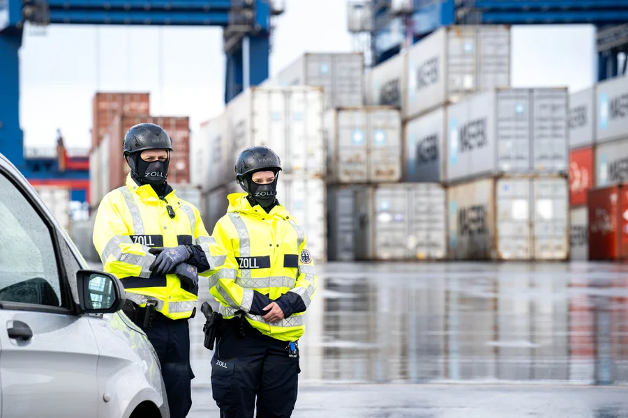 Zwei Polizist_innen stehen in einem Hafen vor Containern.