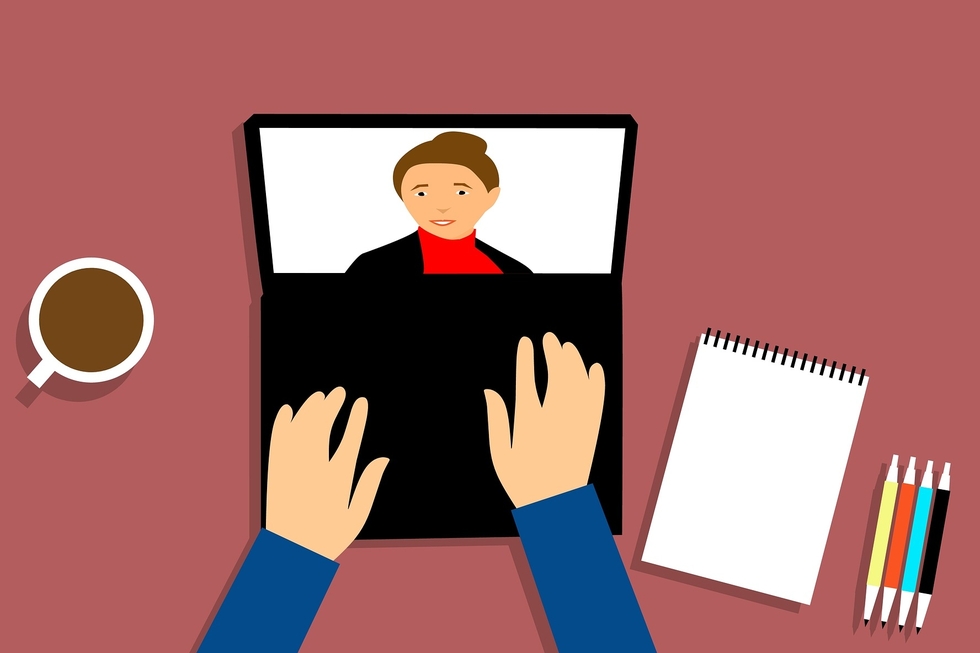 Zwei Hande liegen auf der Tastatur eines aufgeklappten Laptops. Auf dem Bildschirm des Laptops befindet sich eine Person mit hochgebundenen Haaren, rotem Kragenpullover und einer schwarzen Jacke.