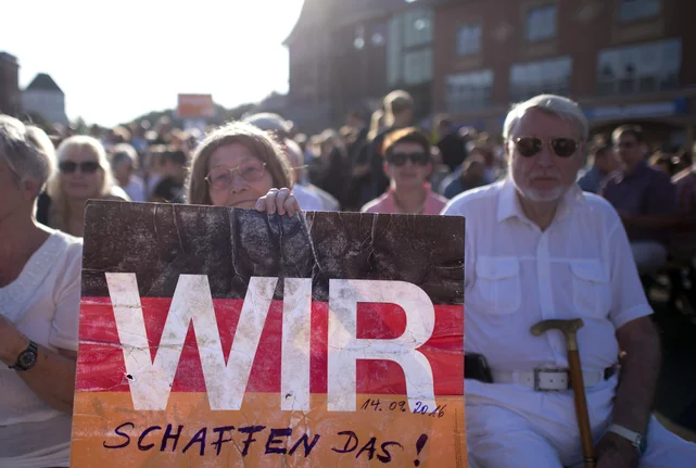 Menschen halten ein Schild mit der Aufschrift "Wir schaffen das!". Das Schild hat die Farben der Deutschlandflagge.