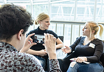 Eine Frau macht mit ihrem Smartphone ein Foto von zwei Frauen auf Sesseln, die sich unterhalten.