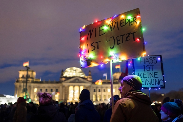 ein Foto von einer Demo vor dem Bundestag in Berlin. Es ist leicht dunkel und auf den Plaketen stehen "Nie wieder ist jetzt" und "Hass ist keine Meinung". Geschmückt wurden diese mit bunten Lichterketten. Im Vordergrund sind zwei Personen mit Mützen und dicken Winterjacken zu sehen.