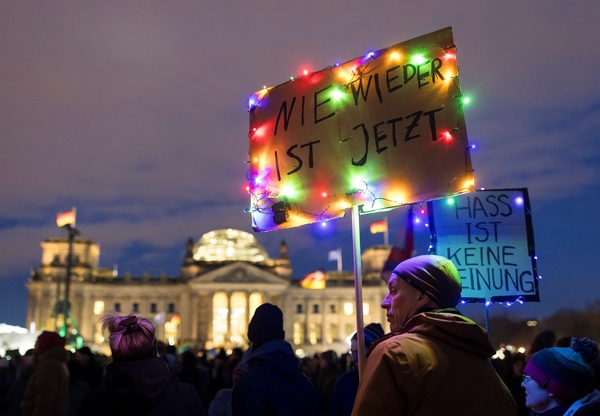 ein Foto von einer Demo vor dem Bundestag in Berlin. Es ist leicht dunkel und auf den Plaketen stehen "Nie wieder ist jetzt" und "Hass ist keine Meinung". Geschmückt wurden diese mit bunten Lichterketten. Im Vordergrund sind zwei Personen mit Mützen und dicken Winterjacken zu sehen.