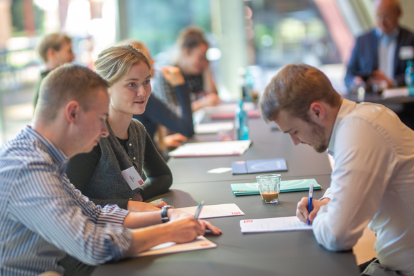 Foto: Mehrere junge Erwachsene sitzen an einem Tisch und notieren sich mit Stiften etwas auf den vor ihnen liegenden Blöcken.
