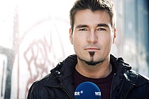 Portrait von Till Opitz, der mit einem blauen WDR-Mikrofon vor einer Wand steht.