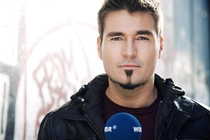 Portrait von Till Opitz, der mit einem blauen WDR-Mikrofon vor einer Wand steht.