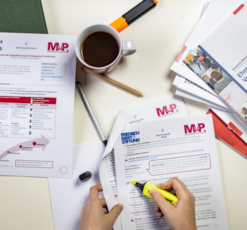 Ein Tisch mit Materialien der MuP-Praxishilfe. Ein Person markiert einen Absatz in einem Dokument.