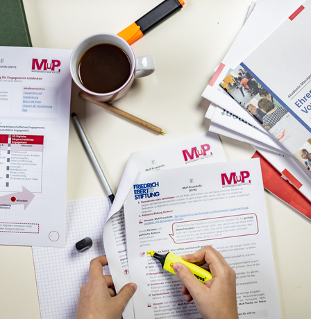 Ein Tisch mit Materialien der MuP-Praxishilfe. Ein Person markiert einen Absatz in einem Dokument.
