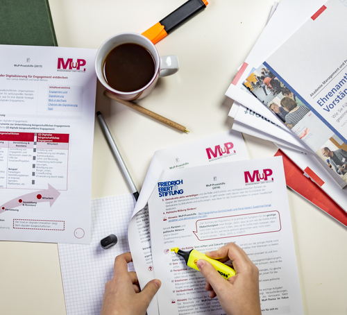 Ein Tisch mit Materialien der MuP-Praxishilfe. Ein Person markiert einen Absatz in einem Dokument.