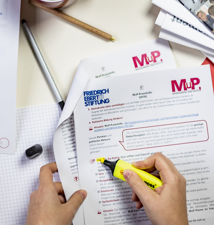 Ein Tisch mit Materialien der MuP-Praxishilfe. Ein Person markiert einen Absatz in einem Dokument.