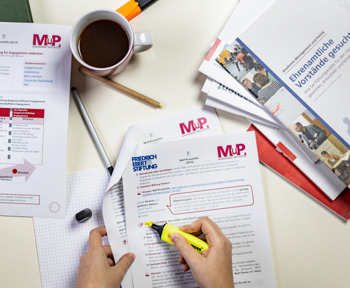 Ein Tisch mit Materialien der MuP-Praxishilfe. Ein Person markiert einen Absatz in einem Dokument.