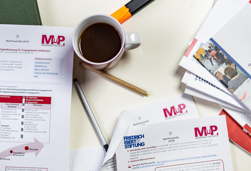 Ein Tisch mit Materialien der MuP-Praxishilfe. Ein Person markiert einen Absatz in einem Dokument.