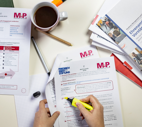 Ein Tisch mit Materialien der MuP-Praxishilfe. Ein Person markiert einen Absatz in einem Dokument.