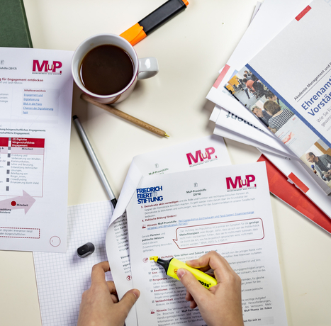 Ein Tisch mit Materialien der MuP-Praxishilfe. Ein Person markiert einen Absatz in einem Dokument.