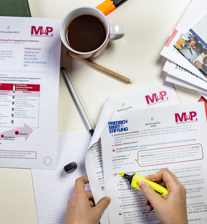 Ein Tisch mit Materialien der MuP-Praxishilfe. Ein Person markiert einen Absatz in einem Dokument.