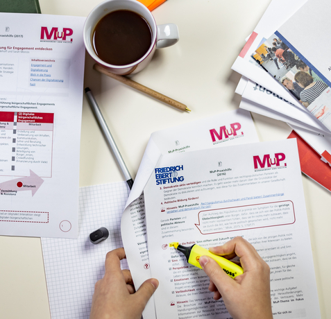 Ein Tisch mit Materialien der MuP-Praxishilfe. Ein Person markiert einen Absatz in einem Dokument.