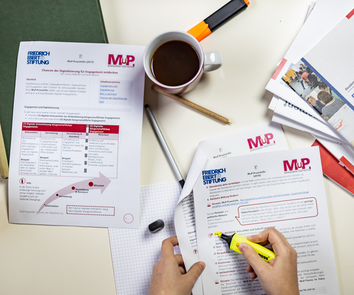 Ein Tisch mit Materialien der MuP-Praxishilfe. Ein Person markiert einen Absatz in einem Dokument.