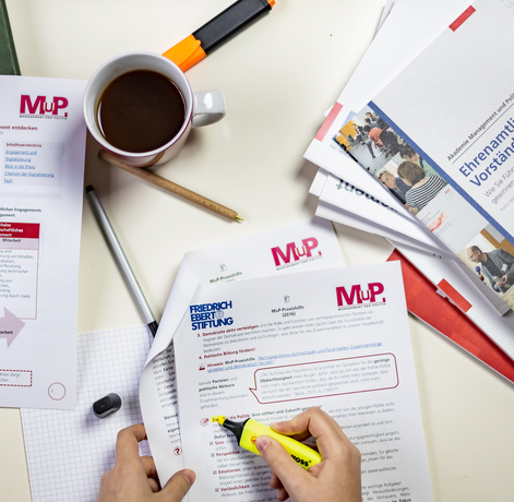 Ein Tisch mit Materialien der MuP-Praxishilfe. Ein Person markiert einen Absatz in einem Dokument.