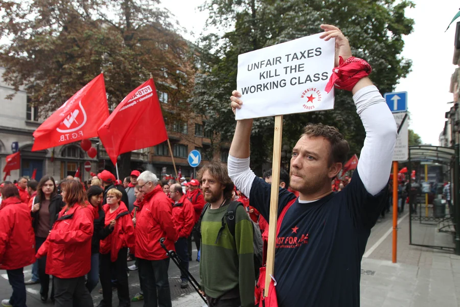 Ein Demonstrant hält ein Plakat mit der Forderung nach gerechten Steuern während einer Demonstration durch Brüssel. Weitere Demonstrant_innen halten rote Fahnen und Banner.