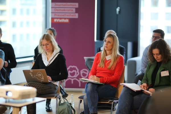 Foto: Mehrere Persone sitzen in einem Workshop auf Stühlen hören zu.